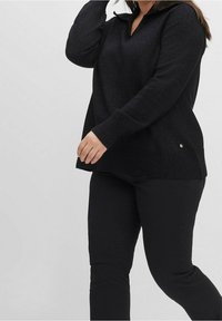 Maglione in maglia nera con scollo a V e orlo a costine, dotato di maniche lunghe e un piccolo dettaglio del logo di lato. Abbinato a pantaloni neri.