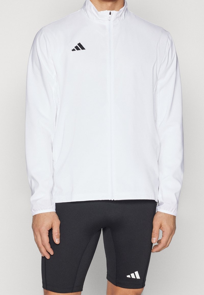 Veste de survêtement blanche avec fermeture éclair intégrale, col montant, manches longues et logo Adidas noir. Associée à un short noir ajusté avec logo blanc.