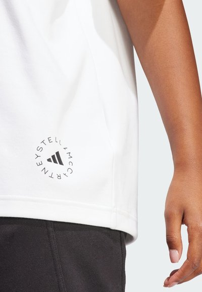 Tricou alb din bumbac cu un logo rotund negru în partea stângă jos; mâneci scurte și o croială lejeră vizibile aproape de o mâna și o pereche de pantaloni negri.