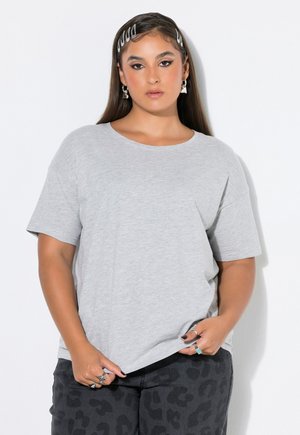 Frau mit langen Haaren, die ein hellgraues T-Shirt und dunkle Hose mit Leopardenmuster trägt, hält den Saum ihres Shirts mit mehreren Ringen an den Fingern.