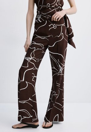 Femeie care poartă pantaloni largi, de culoare maro închis, cu modele abstracte albe, asortați cu un top similar și sandale negre cu talpă joasă.