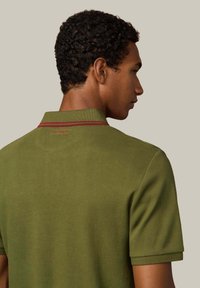 Zelené tričko polo s textúrovanou látkou, s klasickým golierom a červeným pruhom pozdĺž výstrihu. Jemné logo na zadnej strane.