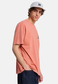 Joven con gorra gris claro, camiseta holgada de color salmón con un pequeño gráfico y vaqueros azul oscuro, de pie con una mano en el bolsillo.