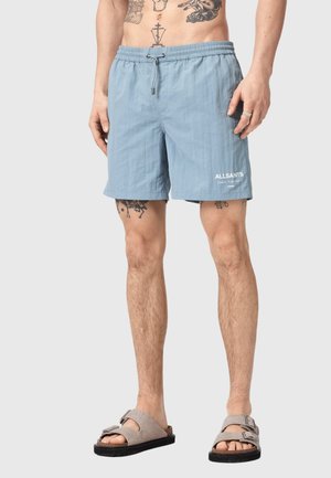 Mężczyzna w jasnoniebieskich szortach z sznurkiem i logo "ALLSAINTS" na lewej nodze oraz w beżowych sandałach na klamry, stojący z widocznymi tatuażami na torsie i nogach.