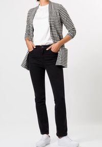 Persona con jeans negros, zapatillas blancas, blusa blanca y chaqueta de cuadros en blanco y negro, con las manos en los bolsillos.