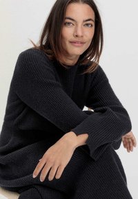 Strickkleid in Schwarz mit gerippter Textur, langen Ärmeln und rundem Ausschnitt. Lockere Passform mit offenen Details an den Ärmelsäumen.