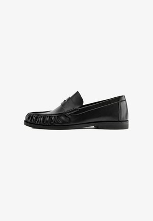 Mocassin en cuir noir avec des détails froncés autour de l’orteil, un petit talon et une bride classique, présenté de profil sur fond blanc.