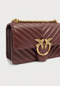 Δερμάτινη τσάντα χιαστί σε βουργουνδί χρώμα με υφή καπιτονέ σε σχέδιο ψαροκόκαλο, μεταλλικά στοιχεία σε χρυσό τόνο και κούμπωμα με μοτίβο πουλιού. Περιλαμβάνεται λουρί αλυσίδα.