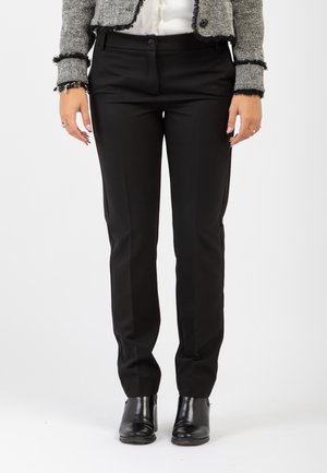 Pantaloni neri con una vestibilità sartoriale, caratterizzati da sottili pieghe frontali e tasche laterali, realizzati in un tessuto morbido. Indossati con stivali alla caviglia.