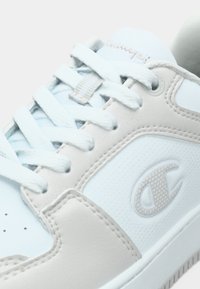 Αθλητικό παπούτσι σε ανοιχτό μπλε και μπεζ με υφή στην επιφάνεια, στρογγυλή μύτη, δετά κορδόνια και λογότυπο στην πλευρά. Κατασκευασμένο από ομαλά υλικά.