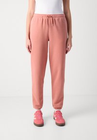 Vrouw in zalmkleurige sweatpants met trekkoord, wit topje en bijpassende roze sneakers, staande tegen een effen achtergrond.