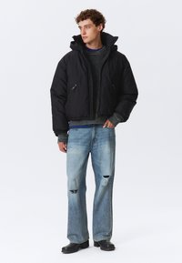 Sort cropped puffer jakke med hætte, to lynlåsede lommer og ribbede manchetter, parret med slidte lyseblå jeans og sorte støvler.