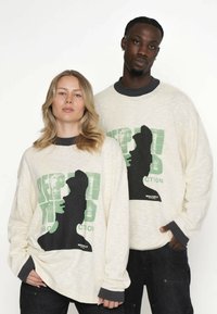 Kremowe bluzy z nadrukami w czerni i zieleni, z dekoltem w serek i kontrastowymi mankietami, noszone przez dwóch modeli.