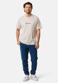 Beige bomulds t-shirt med "Slade Studios" trykt i sort, parret med blå jeans og hvide sneakers. Lige snit med korte ærmer.