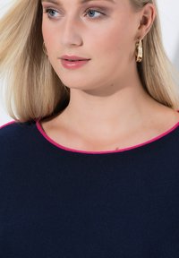 Blonde Frau mit blauen Augen, die goldene Creolen trägt und ein marineblaues Oberteil mit leuchtend pinkem Saum am Ausschnitt.
