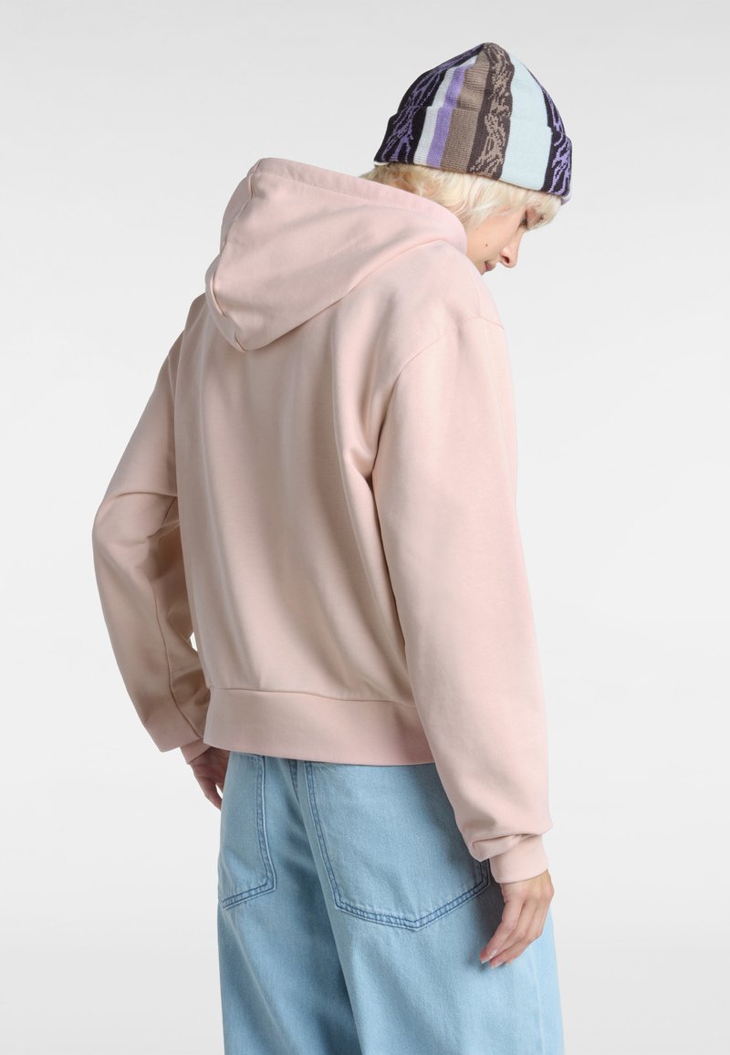 Felpa rosa con cappuccio, dal taglio morbido, con polsini e orlo a coste. Indossata con pantaloni di denim azzurro chiaro. La modella porta un berretto a fantasia.