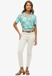 Krótka turkusowa koszula z tropikalnym nadrukiem, zestawiona z białymi dopasowanymi spodniami i brązowymi sandałami na obcasie. Stylizację dopełnia brązowy pasek.