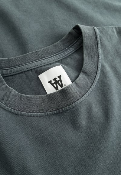 T-shirt in cotone grigio scuro con scollo rotondo, caratterizzato da un orlo cucito e un'etichetta in tessuto con la dicitura "AW". Texture liscia e design semplice.