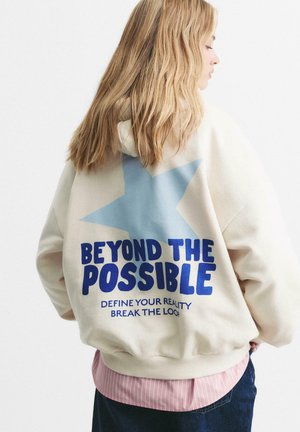 Person with long blonde hair wearing a cream hoodie with a blue star and the text "BEYOND THE POSSIBLE, DEFINE YOUR REALITY, BREAK THE LOOP."