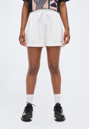 Άτομο που φοράει λευκά σορτς πλέγμα με κορδόνια και μαύρα αθλητικά παπούτσια με λευκές κάλτσες, στέκεται σε απλό φόντο.