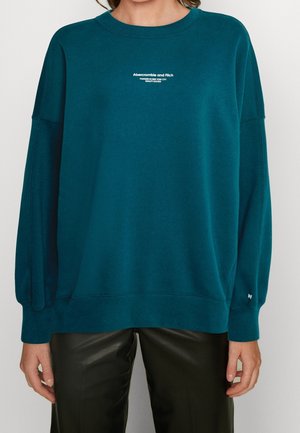 Person, die ein türkisfarbenes Abercrombie and Fitch Crewneck-Sweatshirt und schwarze Lederhosen trägt, steht vor einem einfarbigen hellen Hintergrund.