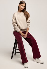 Beżowa bluza z napisem „LEE COOPER” wytłoczonym na biało, w połączeniu z bordowymi szerokimi spodniami ze sztruksu i białymi sneakersami, siedząca na czarnym stołku.