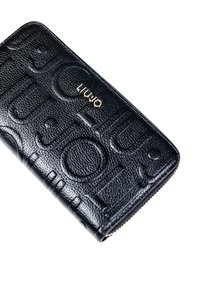 Cartera negra texturizada con lettering en relieve y logotipo dorado. Presenta un cierre con cremallera y un diseño elegante, adecuado para el uso diario.