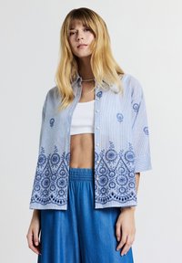 Frau mit langen blonden Haaren, die ein hellblaues besticktes Hemd �über einem weißen Cropped-Top und lockeren blauen Hosen trägt, vor einem einfarbigen Hintergrund stehend.