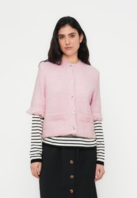 Ροζ ζακέτα με κοντά μανίκια και χνουδωτή υφή, ασημένια κουμπιά και δύο μπροστινές τσέπες· φορεμένη πάνω από μα ύρο-άσπρο ριγέ μακρυμάνικο μπλουζάκι.