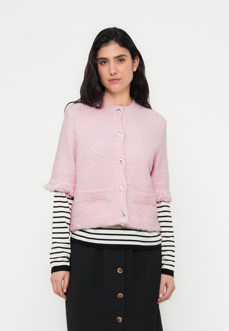 Ροζ ζακέτα με κοντά μανίκια και χνουδωτή υφή, ασημένια κουμπιά και δύο μπροστινές τσέπες· φορεμένη πάνω από μαύρο-άσπρο ριγέ μακρυμάνικο μπλουζάκι.