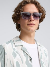 Grijze transparante zonnebril met donkere lenzen, gedragen met een licht gekleed shirt met groene en witte abstracte ontwerpen.