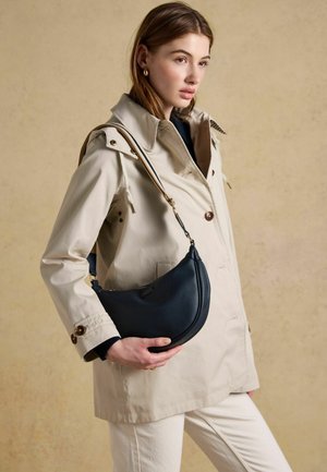 Noor naine beežis trenchmantlis, hoides musta poolkuu-kujulist nahast käekotti triibulise õlarihmaga neutraalsel taustal.