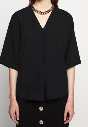 Kvinde iført en sort V-hals bluse med albuelange ærmer og en chunky guldkæde som halskæde.