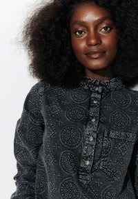 Chemise noire à motifs présentant des designs complexes, un col froncé et une fermeture à boutons sur le devant. Il y a une poche poitrine avec un motif similaire.