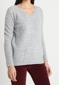 Pull à col en V, à manches longues en gris clair, en tissu tricoté texturé avec un ourlet et des poignets côtelés. Associé à un pantalon bordeaux.