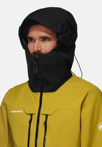 Giacca impermeabile gialla con cappuccio nero, dotata di un colletto alto, chiusura frontale con cerniera e due tasche con cerniera. Texture liscia, minimalismo del branding.