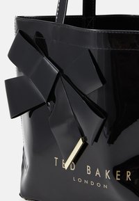 Mustassa kiiltävässä olkalaukussa on suuri musta nauhakoriste, kohokuvioitu "TED BAKER LONDON" -logo ja sileä pinta. Kahvat helppoa kantamista varten.