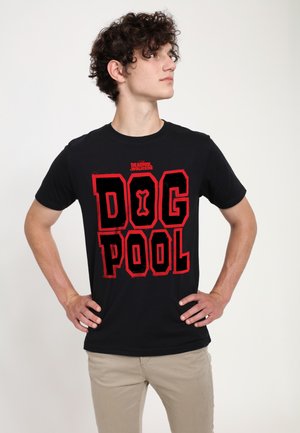 Jonge man met krullend haar, staand met handen in de zij, die een zwart Deadpool Wolverine T-shirt draagt met rode tekst "DOG POOL".