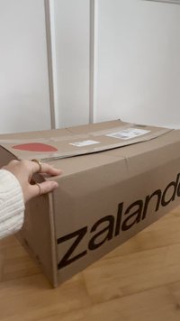 Ruda kartoninė dėžutė su juodu spaudiniu logotipu. Dėžutė turi plokščią viršų, švelnias raukšles ir šoną puošiantį raudoną apskritą lipduką.