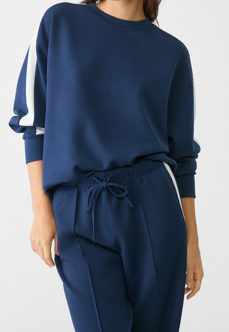 Felpa blu navy con strisce bianche ai lati, con collo rotondo e maniche lunghe, abbinata a pantaloni della tuta blu navy con vita elastica.