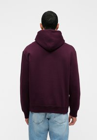 Hanorac cu glugă de culoare bordo, cu o croială lejeră, dintr-un material textil gros, cu manșete din tricot, purtat cu blugi de culoare bleu deschis. Design simplu, de o culoare solidă.