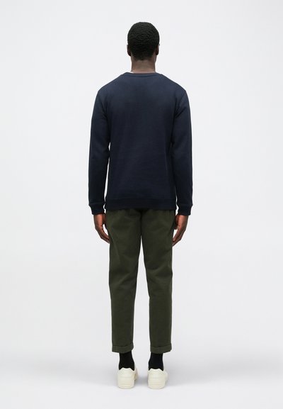 Marine crew neck sweatshirt, bomuldsblanding, afslappet pasform; parret med olivengrønne tætsiddende bukser; hvide sneakers; ensfarvet baggrund.