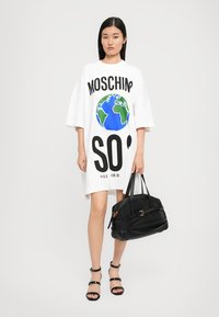 Weißes, übergroßes Kleid mit "MOSCHINO" und Erdgrafikdesign. Schwarze Handtasche mit Reißverschlüssen und Riemen. Die Model trägt schwarze Sandalen mit Riemchen.