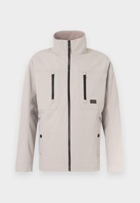 Veste beige clair avec un col haut, deux poches poitrine zippées et des accents de fermeture éclair noirs. Texture lisse et design épuré.