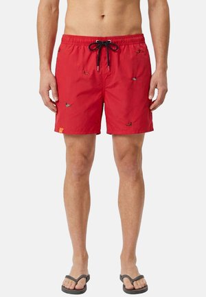 Uomo che indossa pantaloncini da bagno rossi con piccoli cani ricamati, coulisse nera e infradito nere, in piedi su sfondo bianco.