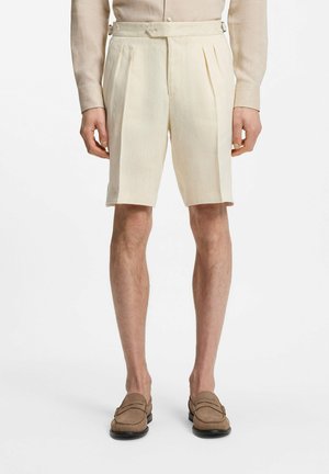 Beige plissierte Shorts mit seitlichen Schnallendetails, getragen mit passendem beigen Hemd und braunen Loafers an einem männlichen Modell.