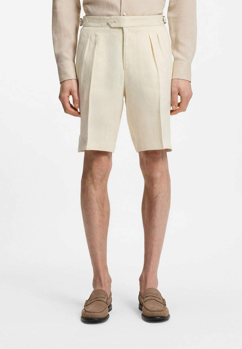 Shorts beiges plissés avec des détails de boucle latérale portés avec une chemise beige assortie et des mocassins marron sur un modèle masculin.