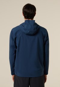 Hombre de espaldas llevando una chaqueta azul marino con capucha y mangas largas, y pantalones negros, sobre un fondo beige liso.