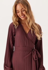 Robe cache-cœur couleur prune avec des manches longues et une texture plissée. Comprend une ceinture à nouer à la taille et un décolleté en V profond.