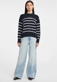 Femeie în picioare, purtând un pulover cu dungi negre și albe de la Guess Jeans, blugi largi de culoare albastru deschis și pantofi cu vârf ascuțit negri, pe un fundal alb.